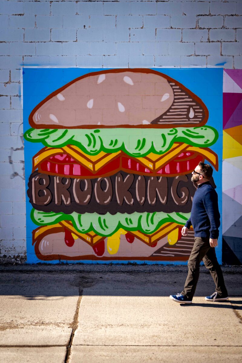 man walking in front of an Urban Canvas mural featuring a hamburger that has Brookings written in it.