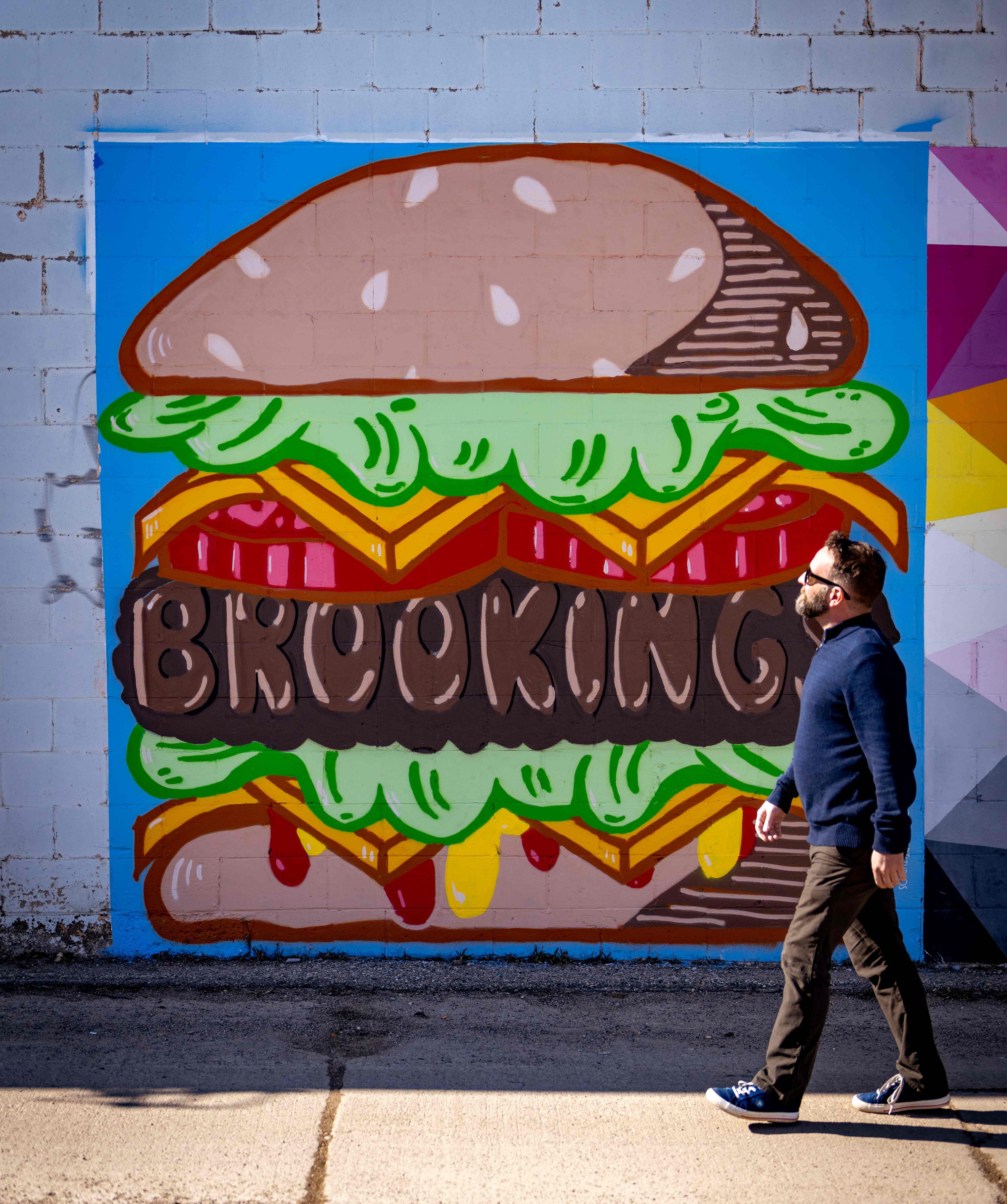 man walking in front of an Urban Canvas mural featuring a hamburger that has Brookings written in it.