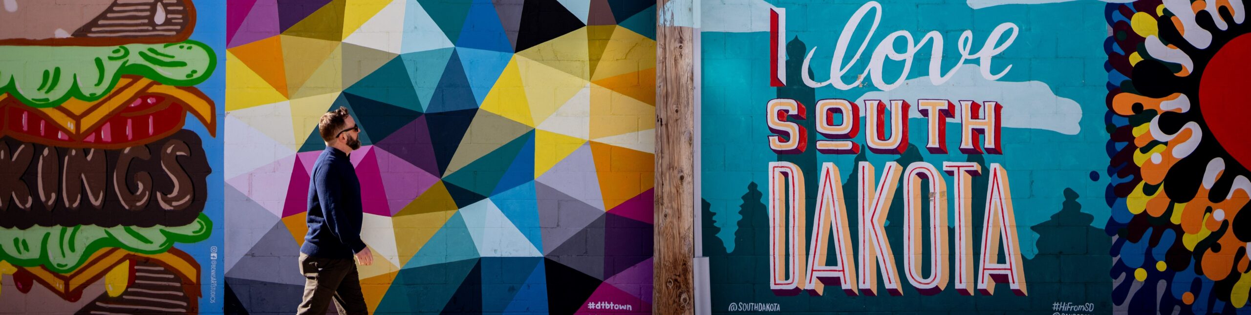 A colorful mural scene on a city wall featuring three distinct sections. On the left, a giant vibrant hamburger mural has the word 'BROOKINGS' written across the patty. In the middle, a geometric mural with multicolored triangles in shades of blue, yellow, pink, and gray creates a lively pattern. On the right, a mural displays the phrase 'I love SOUTH DAKOTA' in bold red and white letters against a blue background, with a large red heart and colorful abstract shapes surrounding it. A man in a blue sweater and sunglasses walks past the murals, looking up at the artwork