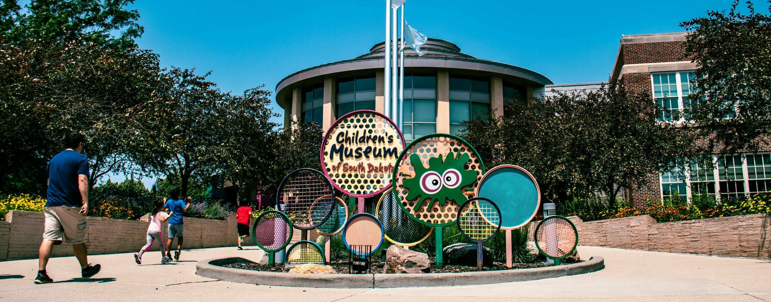 Children's Museum of South Dakota entrance featuring a vibrant, playful sign surrounded by colorful decor and greenery. The modern building and a bright blue sky provide a welcoming backdrop, with families walking toward the museum.