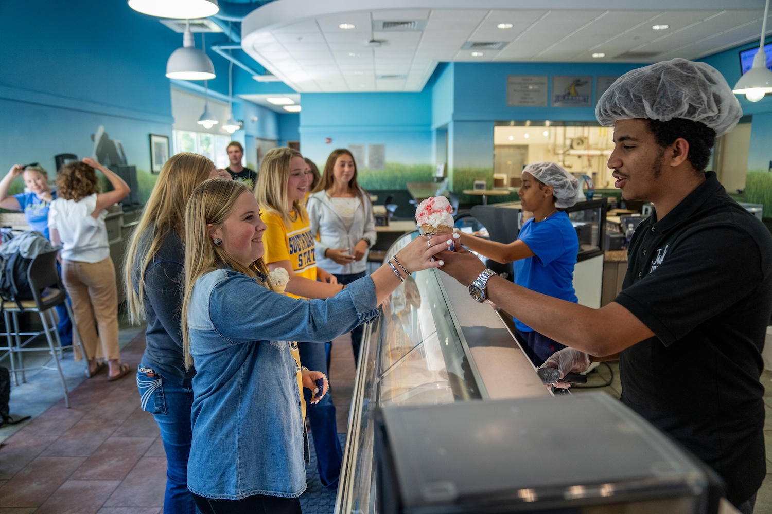 A student is handed ice cream by an employee at the SDSU Dairy Bar. In the background there are students in line waiting amidst blue painted walls.