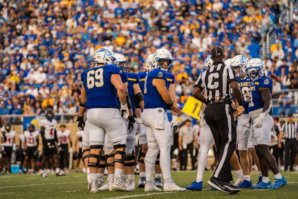 Jackrabbit football players gather with the crowd to their back. In between plays, they players look to be in high spirits. A ref is also gathered with them.