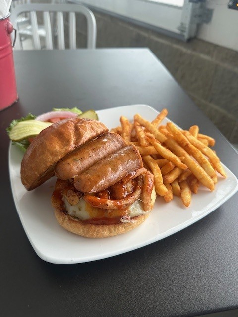 A burger with a sausage link served on top with a red sauce. Served with fries